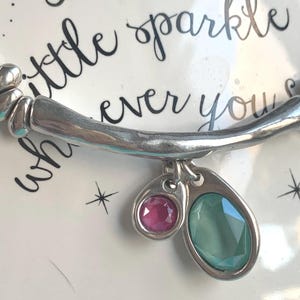 May include: Silver-toned bracelet with two charms, one with a pink gemstone and the other with a green gemstone. The bracelet is set against a white background with the words "little sparkle ever you" in cursive.