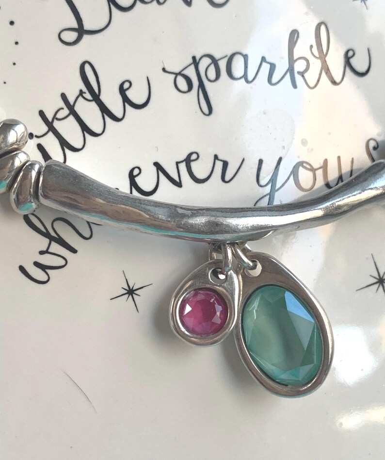 May include: Silver-toned bracelet with two charms, one with a pink gemstone and the other with a green gemstone. The bracelet is set against a white background with the words "little sparkle ever you" in cursive.
