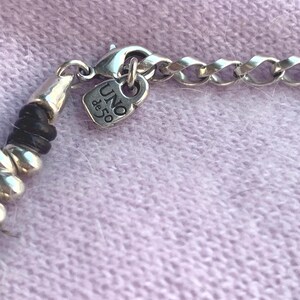 May include: Close-up of a silver necklace with a chain link design. The necklace features a small, heart-shaped charm with the text "UNO de 50". The necklace is on a light purple surface.