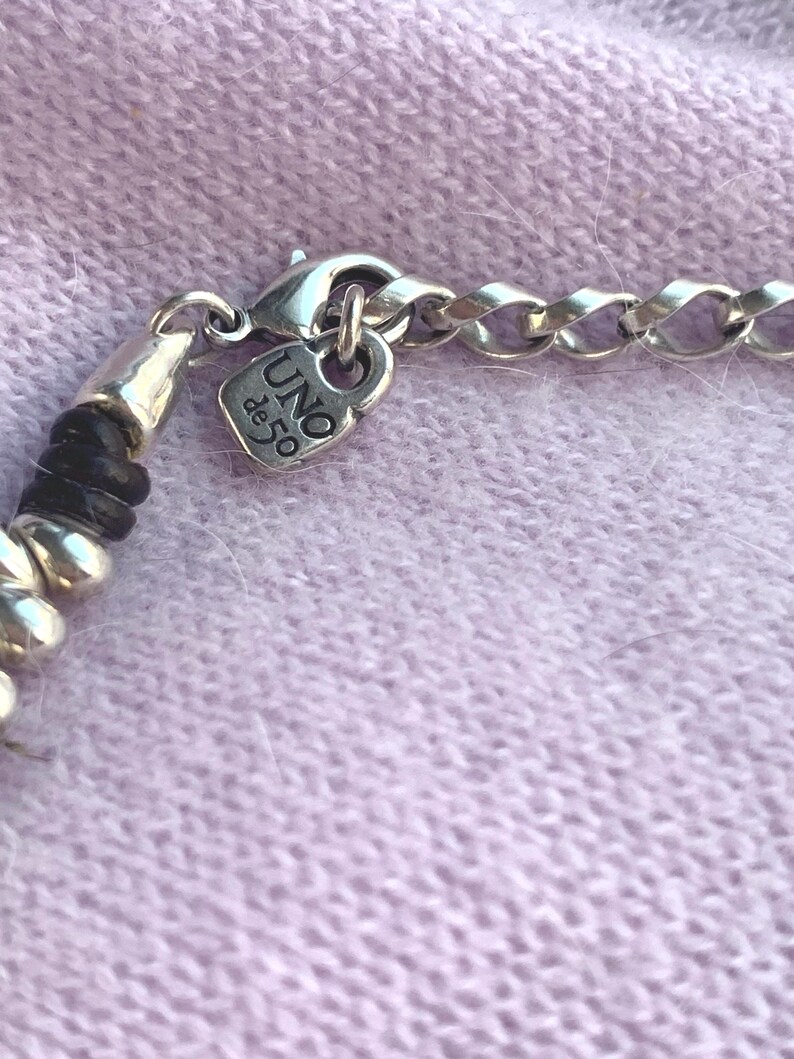 May include: Close-up of a silver necklace with a chain link design. The necklace features a small, heart-shaped charm with the text "UNO de 50". The necklace is on a light purple surface.