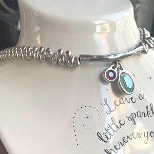 May include: A silver necklace with a beaded design and two gemstone charms, one red and one green. The necklace is displayed on a white mannequin. The text "Leave a little sparkle wherever you go" is visible.