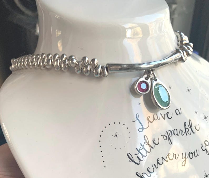May include: A silver necklace with a beaded design and two gemstone charms, one red and one green. The necklace is displayed on a white mannequin. The text "Leave a little sparkle wherever you go" is visible.