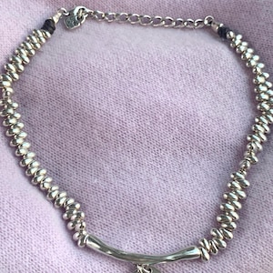 May include: Silver-toned necklace with a beaded design and a curved bar pendant. The pendant features a small pink and a larger teal-colored gemstone. The necklace is displayed on a light purple fabric.