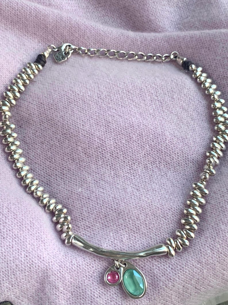 May include: Silver-toned necklace with a beaded design and a curved bar pendant. The pendant features a small pink and a larger teal-colored gemstone. The necklace is displayed on a light purple fabric.