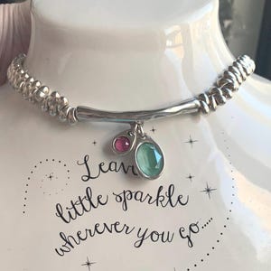 May include: Silver-toned necklace with a beaded band and a curved bar pendant. The pendant features a small pink and a larger green gemstone. The text "Leave little sparkle wherever you go..." is visible.