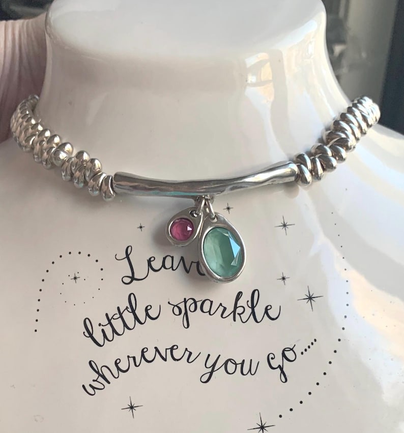 May include: Silver-toned necklace with a beaded band and a curved bar pendant. The pendant features a small pink and a larger green gemstone. The text "Leave little sparkle wherever you go..." is visible.