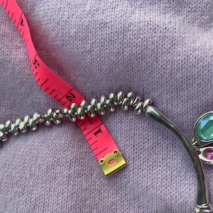 May include: Silver-toned necklace with a beaded design and a curved pendant featuring a large oval teal gemstone and a smaller pink gemstone. A pink measuring tape is placed across the necklace.