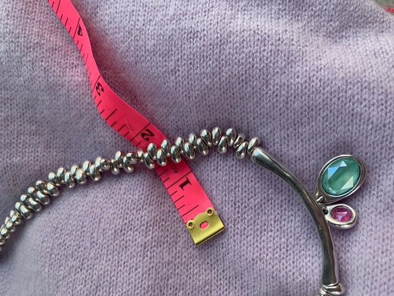 May include: Silver-toned necklace with a beaded design and a curved pendant featuring a large oval teal gemstone and a smaller pink gemstone. A pink measuring tape is placed across the necklace.