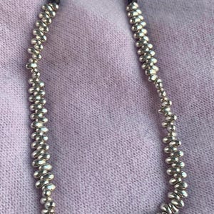 May include: Silver-toned necklace with a beaded design and a curved bar pendant. The pendant features a green oval-shaped stone and a small pink stone. The necklace is displayed on a light purple fabric.