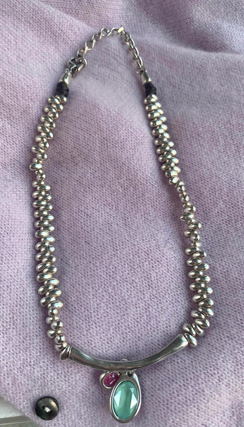 May include: Silver-toned necklace with a beaded design and a curved bar pendant. The pendant features a green oval-shaped stone and a small pink stone. The necklace is displayed on a light purple fabric.