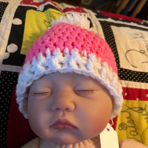 May include: A sleeping baby doll wearing a pink and white crocheted hat with a white pom-pom on top.