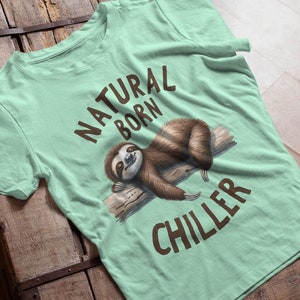 Könnte beinhalten: Ein hellgrünes T-Shirt mit einem braunen Faultier-Motiv und dem Text "NATURAL BORN CHILLER".