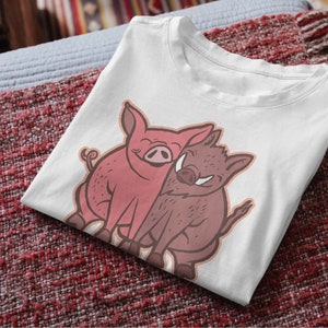 Pode incluir: Camiseta branca com um gráfico de um porco rosa e um javali marrom se abraçando.