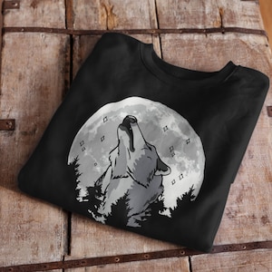 Könnte beinhalten: Schwarzes Sweatshirt mit einem weißen Wolf, der zum Mond heult. Der Mond ist voll und hat eine weiße, strukturierte Oberfläche. Der Wolf ist im Vordergrund und der Mond dahinter. Der Wolf heult zum Mond und hat eine weiße, strukturierte Oberfläche. Der Wolf ist von Bäumen umgeben.