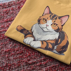 Può includere: Una maglietta gialla con un'immagine di un gatto calico con pelliccia marrone, bianca e arancione.
