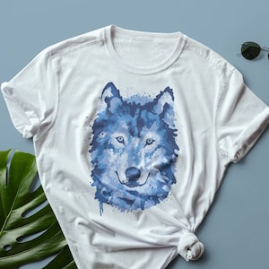 Könnte beinhalten: Weißes T-Shirt mit einem blauen Aquarell-Wolf-Design. Das Gesicht des Wolfes ist mittig auf dem Shirt platziert und hat eine detaillierte, realistische Optik.