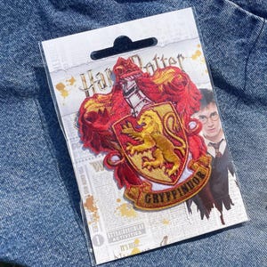 Può includere: Una toppa di Grifondoro di Harry Potter su una carta, con lo stemma di Grifondoro con un leone, colori rosso e oro e la parola "Grifondoro". La carta mostra anche un'immagine di Harry Potter. La toppa e la carta sono su una superficie di denim.
