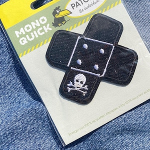 Puede incluir: Un parche termoadhesivo negro y blanco con un diseño de calavera y huesos cruzados sobre un vendaje blanco. El parche está etiquetado como "MONO QUICK PATCHES Be individual".