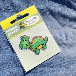 Könnte beinhalten: Eine Packung Bügelflicken mit einem grünen Dinosaurier-Design. Der Aufnäher befindet sich auf einem weißen Hintergrund mit dem Text "MONO QUICK" und "PATCHES Be individual". Die Packung liegt auf einer blauen Jeansoberfläche.