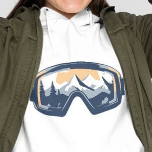 Op de afbeelding: Witte hoodie met een afbeelding van blauwe skibrillen met een berglandschap in de lenzen. De hoodie wordt gedragen door een persoon met een zwarte beanie.