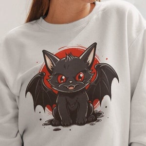 Könnte beinhalten: Weißer Pullover mit einer schwarzen Katze mit roten Augen und Fledermausflügeln. Die Katze befindet sich vor einem roten Kreis.