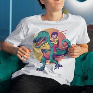 Peut inclure: Un t-shirt blanc avec un motif représentant un personnage de dessin animé chevauchant un dinosaure. Le dinosaure est vert et a une selle rouge et jaune. Le personnage porte une chemise bleue et un chapeau rouge.