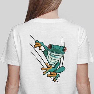 Peut inclure: T-shirt blanc avec une grenouille verte qui sort d'un arbre. La grenouille a des pieds orange et des yeux rouges.