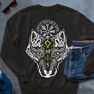 Könnte beinhalten: Schwarzes Sweatshirt mit einem weißen Wolfskopf-Motiv und einem gelben Kompass-Symbol. Die Augen des Wolfes sind gelb und der Kompass trägt den Text "Vegvísir" um sich herum.