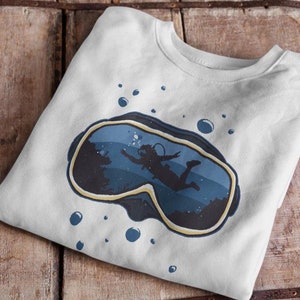 Può includere: Felpa bianca con una grafica di una maschera da subacquea blu e gialla. La maschera mostra un subacqueo che nuota sott'acqua con delle bolle.