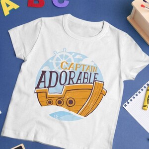 Puede incluir: Camiseta blanca con un gráfico de un barco amarillo con un volante y el texto "Captain Adorable".