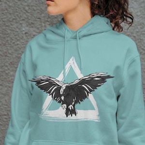 Può includere: Una felpa blu chiaro con un'immagine di un corvo nero all'interno di un triangolo bianco. Il corvo sta volando con le ali spiegate.