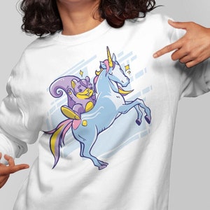 Op de afbeelding: Wit sweatshirt met een afbeelding van een paarse eekhoorn die op een blauwe eenhoorn rijdt met een roze hoorn en manen. De eenhoorn staat op zijn achterpoten.