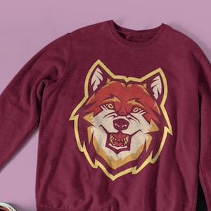 Könnte beinhalten: Ein maroonfarbener Pullover mit einem Aufdruck eines roten und gelben Wolfskopfes. Der Wolf hat einen grimmigen Ausdruck und ist von einem gelben Umriss umgeben.