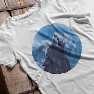 Könnte beinhalten: Weißes T-Shirt mit einem runden Motiv eines Wolfes, der zum Mond heult. Der Mond ist blau und weiß mit Wolken.