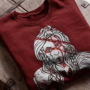 Könnte beinhalten: Burgunderfarbener Pullover mit einem weißen Grafikdruck einer Frau mit langen Haaren und einem roten Strich über dem Gesicht.