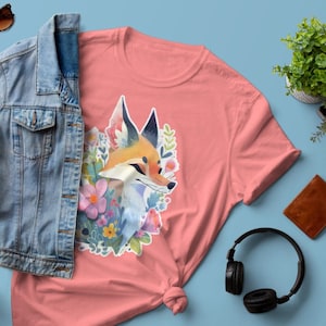 Könnte beinhalten: Ein pinkfarbenes T-Shirt mit einem Aufdruck eines Fuchses, der von Blumen umgeben ist. Der Fuchs hat orangefarbenes Fell und eine weiße Brust. Die Blumen sind in Rosa-, Lila- und Gelbtönen gehalten. Das T-Shirt liegt auf einer blauen Oberfläche mit einer Jeansjacke, einer Sonnenbrille, einem Notizbuch, einer Pflanze und Kopfhörern.