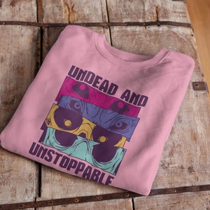 Könnte beinhalten: Rosafarbener Pullover mit einem Grafikdruck von bunten Schädeln und dem Text "Undead and Unstoppable".