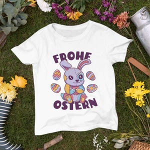 Puede incluir: Camiseta blanca con un conejito de dibujos animados morado y rosa que sostiene un huevo de colores. El texto "Frohe Ostern" está impreso en la camiseta.