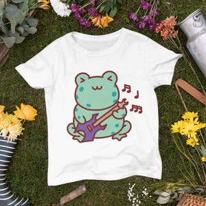 Puede incluir: Camiseta blanca con una rana verde tocando una guitarra eléctrica morada. La rana tiene una expresión feliz y hay notas musicales sobre su cabeza.