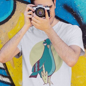 Peut inclure: T-shirt blanc avec un dessin de pigeon de dessin animé. Le pigeon est turquoise et violet, faisant un doigt d'honneur. Le dessin est placé sur un cercle vert clair. Une personne porte le t-shirt et tient un appareil photo.