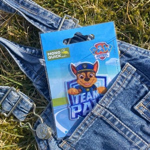 Puede incluir: Un mono de mezclilla azul con un parche de Paw Patrol que presenta un perro de dibujos animados con un sombrero azul y el texto "Team Paw" sobre un fondo blanco. El parche está unido al bolsillo del mono.