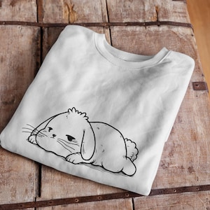 Può includere: Maglietta bianca con un disegno a linea nera di un coniglio dei cartoni animati con gli occhi chiusi e un'espressione triste. Il coniglio è sdraiato di lato.