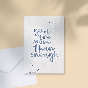 May include: A white greeting card with the handwritten phrase "you are more than enough" in blue ink. The card has blue ink splatters and is next to a white envelope on a beige surface.