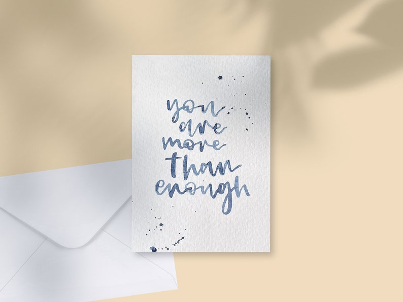 May include: A white greeting card with the handwritten phrase "you are more than enough" in blue ink. The card has blue ink splatters and is next to a white envelope on a beige surface.