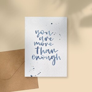 May include: A white watercolour card with the blue script text: "you are more than enough". The card has blue paint splatters and is next to a brown envelope. The background is a light beige colour.