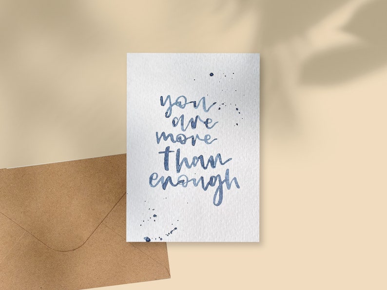 May include: A white watercolour card with the blue script text: "you are more than enough". The card has blue paint splatters and is next to a brown envelope. The background is a light beige colour.