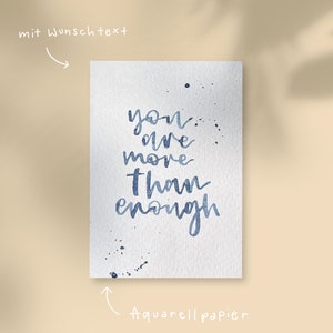 May include: A watercolour painting on textured paper with the words "you are more than enough" in blue script. Splatter details accent the text. The words "mit Wunschtext" and "Aquarellpapier" are also present.