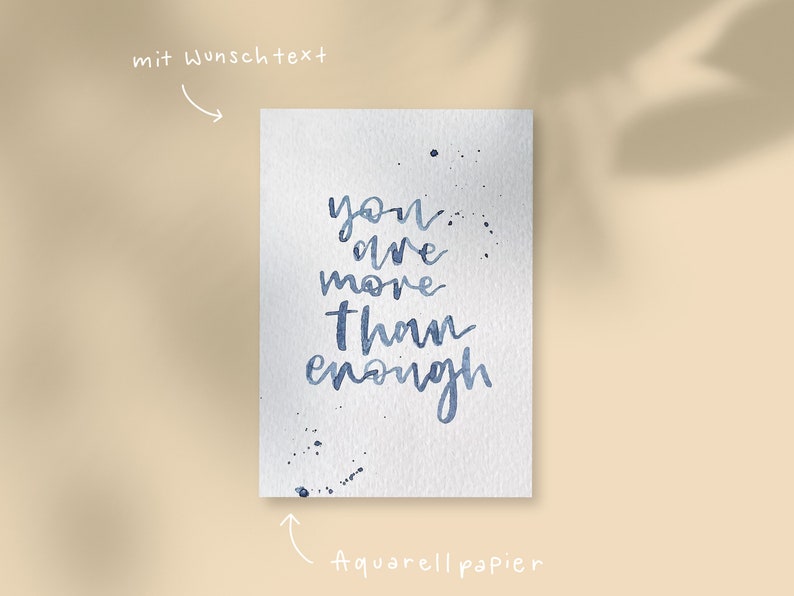May include: A watercolour painting on textured paper with the words "you are more than enough" in blue script. Splatter details accent the text. The words "mit Wunschtext" and "Aquarellpapier" are also present.