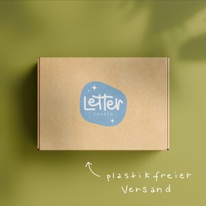 May include: A brown cardboard box with a blue logo reading "Letter Zauber" on a green background. The box has a handwritten arrow pointing to the text "plastic free shipping".