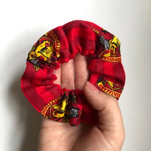 May include: Red and yellow fabric hair scrunchie with a repeated pattern of the Gryffindor house crest from the Harry Potter series. The crest features a lion, a house name, and a diagonal stripe.
